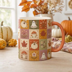 May include: A ceramic mug featuring a patchwork grid design with illustrations of white pumpkins, ghosts, mushrooms, autumn leaves, and wildflowers in shades of orange, green, yellow, and purple. The mug has a solid orange handle and a white interior.
