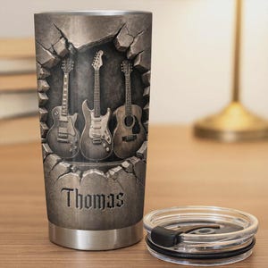 May include: A stainless steel tumbler featuring three guitars emerging from a cracked stone wall design. The name "Thomas" is engraved below the guitars. The tumbler includes a clear lid with a black slider.