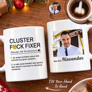 May include: A white ceramic mug featuring black text that reads CLUSTER F*CK FIXER with a definition and a smiley face icon. The mug also displays a photograph of a man in a white shirt and tie with the text See also: Alexander below the image.