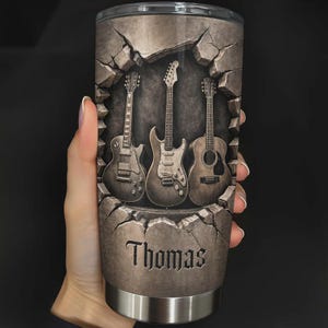 May include: A stainless steel tumbler featuring three guitars set against a cracked stone background. The name "Thomas" is displayed in a decorative font. The design is in shades of gray and brown.