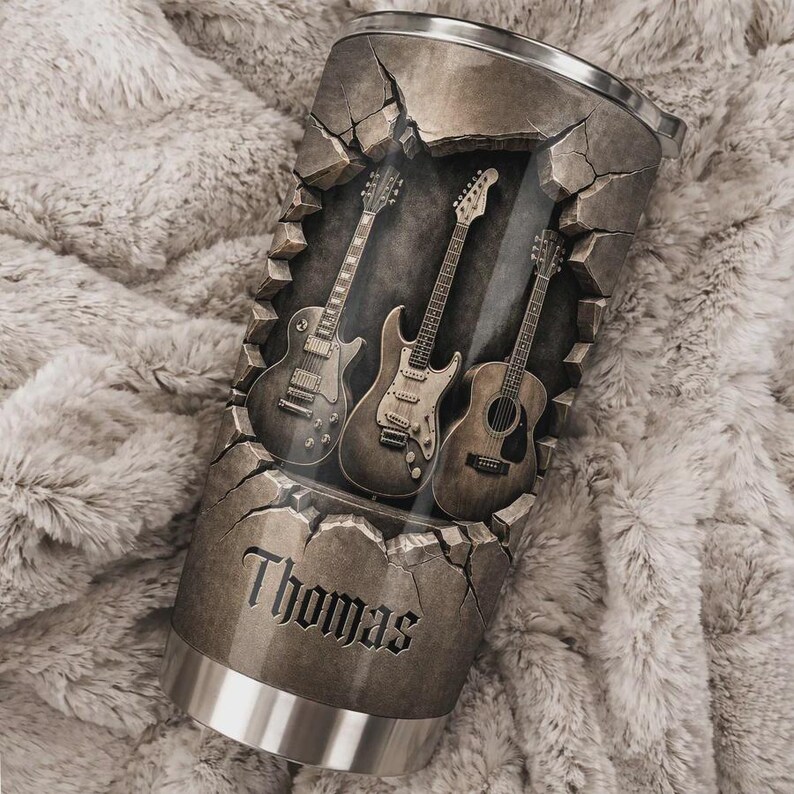 May include: A stainless steel tumbler with a cracked stone design, featuring three guitars. The name "Thomas" is displayed in a gothic font. The tumbler has a silver rim and base, perfect for music lovers.