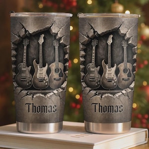 May include: Stainless steel tumbler featuring a design of guitars emerging from a stone wall. The guitars are in shades of gray and brown. The name "Thomas" is printed in a gothic font at the bottom of the tumbler.
