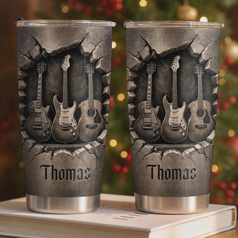 May include: Stainless steel tumbler featuring a design of guitars emerging from a stone wall. The guitars are in shades of gray and brown. The name "Thomas" is printed in a gothic font at the bottom of the tumbler.