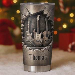 May include: A stainless steel tumbler featuring three guitars emerging from a cracked stone wall design. The name "Thomas" is displayed in a gothic font at the bottom of the tumbler. The background is blurred with Christmas decorations.