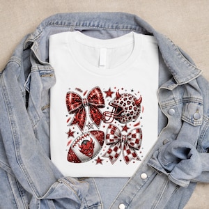 以下が含まれることがあります： 白いTシャツに、2つの大きなリボン、角の生えたフットボールヘルメット、フットボールのグラフィックプリントが施されています。すべて赤と黒のヒョウ柄と小さな赤い悪魔の顔で装飾されており、青いデニムジャケットの下に重ね着されています。