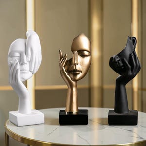 May include: Three abstract face sculptures in white, gold, and black. The white sculpture features hands covering the face. The gold sculpture has a hand supporting the face. The black sculpture has a hand holding the face. All are on black bases.