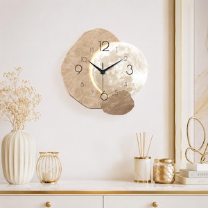 Könnte beinhalten: Eine Wanduhr mit einer unregelmäßigen, strukturierten beigen steinartigen Rückwand und einem kreisförmigen, beleuchteten Zifferblatt im Monddesign. Die Uhr hat schwarze Zeiger, schwarze arabische Ziffern und ein kleines Pendelgewicht.