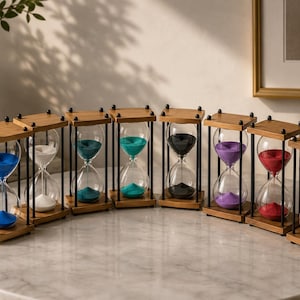 Op de afbeelding: Een verzameling van acht zandlopers opgesteld in een halve cirkel, elk met een houten frame met zwarte metalen steunen en doorzichtige glazen bollen gevuld met gekleurd zand in blauw, wit, groenblauw, turkoois, zwart, paars, rood en beige.