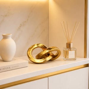 Peut inclure: Un vase en céramique blanc cassé mat, une sculpture décorative en maillons de chaîne métalliques dorés avec une finition texturée, et un flacon diffuseur à roseaux en verre transparent avec des bâtonnets, disposés sur une étagère.