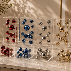 Op de afbeelding: Acht doorzichtige acryl displayboxen met metalen bollen in rood, blauw, zilver en goud. Vier hoge rechthoekige en vier vierkante boxen zijn in twee rijen gerangschikt, elk gevuld met verschillende maten glanzende, reflecterende ballen.