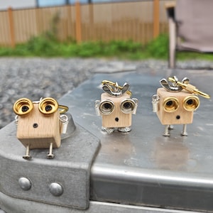 Puede incluir: Tres figuras de robots de madera hechas a mano con detalles en color latón. Cada robot tiene un cuerpo cuadrado de madera, patas de metal y detalles de ojos dorados. Los robots se exhiben sobre una superficie metálica plateada.