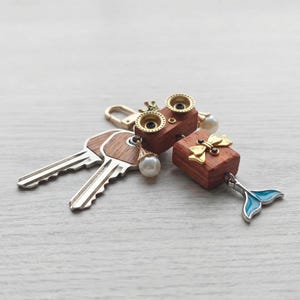 May include: Two keys and two wooden keychains with gold accents and pearl drops. One keychain has a crown design, the other a bow. A blue and silver mermaid tail charm hangs from the second keychain. The keychains are made of wood and metal.
