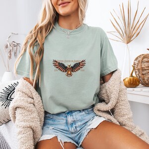 May include: A light green t-shirt featuring a detailed graphic of an eagle with outstretched wings, incorporating the American flag's colors. The text "United States of America" is above the eagle. The shirt is paired with denim shorts.