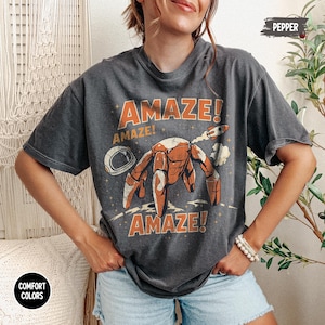 May include: A charcoal gray short-sleeve t-shirt featuring a graphic print of a robotic creature, an astronaut helmet, and a small rocket. The text "AMAZE!" is printed in orange above and below the central illustration, with "PEPPER" in the corner.