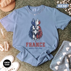 May include: Blue jean colored t-shirt with a graphic of a rooster in red, white, and blue. The word "FRANCE" is printed below the rooster, with "WORLD CUP 2026" underneath. The shirt is surrounded by accessories.
