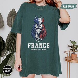 May include: A dark teal t-shirt features a graphic of a rooster in the colors of the French flag. The word "FRANCE" is printed below the rooster, with "WORLD CUP 2026" underneath. The shirt is labeled "Comfort Colors" and "Blue Spruce".