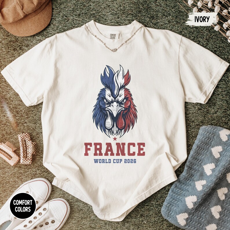 May include: Ivory-colored t-shirt featuring a detailed rooster illustration in red, white, and blue, with the text "FRANCE WORLD CUP 2026". The shirt is part of the Comfort Colors collection.