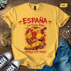 May include: A yellow t-shirt featuring a graphic print with the text "ESPAÑA La Furia Roja" and "WORLD CUP 2026". The design includes a soccer ball, a soccer cleat, and crown icons in red and yellow paint splatter style with a small "DAISY" label.