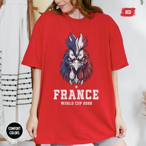 May include: A red t-shirt featuring a detailed graphic of a rooster in the colors of the French flag. The text "FRANCE WORLD CUP 2026" is printed below the rooster. The shirt is a Comfort Colors brand.