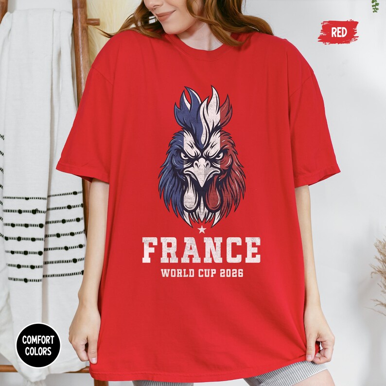 May include: A red t-shirt featuring a detailed graphic of a rooster in the colors of the French flag. The text "FRANCE WORLD CUP 2026" is printed below the rooster. The shirt is a Comfort Colors brand.