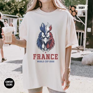 May include: Ivory-colored t-shirt featuring a stylized rooster in the colors of the French flag: blue, white, and red. The word "FRANCE" is printed in red block letters below the rooster, with "WORLD CUP 2026" underneath. The shirt is part of the Comfort Colors collection.
