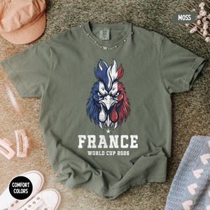 May include: A moss green t-shirt featuring a rooster design in the colors of the French flag: blue, white, and red. The word "FRANCE" is printed below the rooster, along with "WORLD CUP 2026". The shirt has a crew neck and short sleeves.