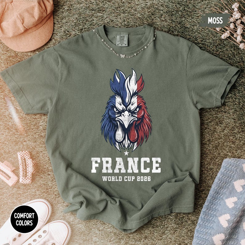 May include: A moss green t-shirt featuring a rooster design in the colors of the French flag: blue, white, and red. The word "FRANCE" is printed below the rooster, along with "WORLD CUP 2026". The shirt has a crew neck and short sleeves.