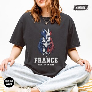 May include: Graphite-colored t-shirt featuring a detailed illustration of a rooster in the colors of the French flag. The text "FRANCE WORLD CUP 2026" is printed below the rooster. The shirt is part of the Comfort Colors collection.
