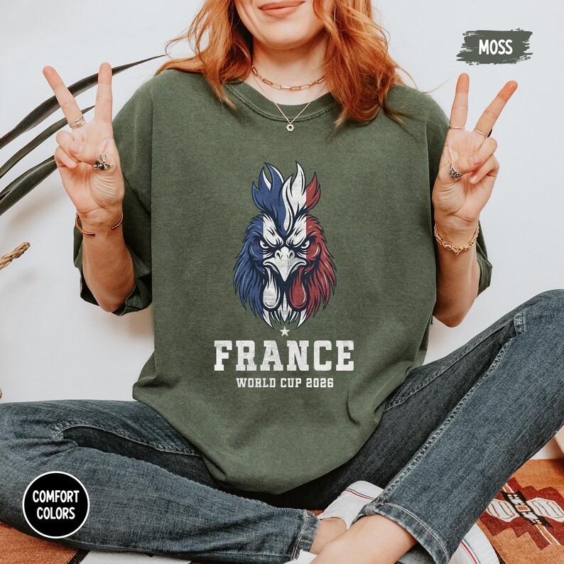 May include: A moss green sweatshirt features a rooster design in the colors of the French flag. The word "FRANCE" is printed below the rooster, with "WORLD CUP 2026" underneath. The sweatshirt is part of the Comfort Colors collection.