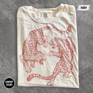 Könnte beinhalten: Ein elfenbeinfarbenes Kurzarm-T-Shirt mit einem roten Grafikdruck von zwei Leoparden in kreisförmiger Anordnung. Das Shirt hat ein Comfort Colors Markenetikett am inneren Halsausschnitt. Der Text IVORY ist oben rechts im Bild aufgedruckt.