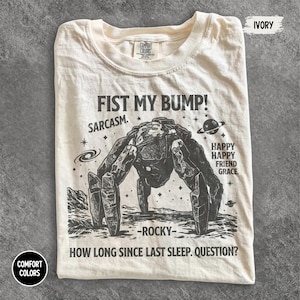 May include: An ivory t-shirt featuring a dark graphic of a rocky, multi-legged creature on a lunar surface. Text includes FIST MY BUMP!, SARCASM., HAPPY HAPPY FRIEND GRACE., -ROCKY-, and HOW LONG SINCE LAST SLEEP. QUESTION? The shirt has a Comfort Colors label.