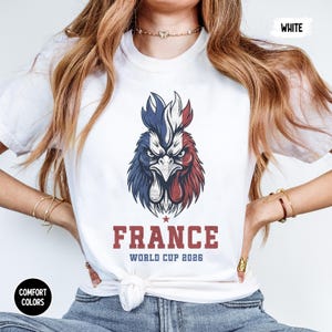 May include: White t-shirt featuring a detailed graphic of a rooster in the colors of the French flag. The word "FRANCE" is printed in a bold, red font, with "WORLD CUP 2026" below. The shirt also has "COMFORT COLORS" and "WHITE" printed on it.