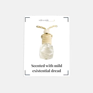 Pode incluir: Purificador de ar de vidro transparente com tampa de madeira e cordão decorativo. O texto no cartão diz "with a wink" e "Scented with mild existential dread."