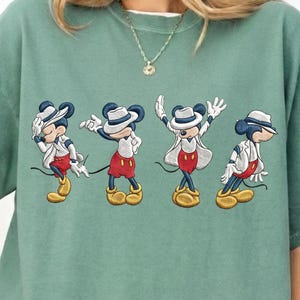May include: Sage green sweatshirt featuring an embroidered design of Mickey Mouse in four different poses. Mickey is wearing a white hat, white jacket, red shorts, and yellow shoes. The design is centered on the front of the shirt.
