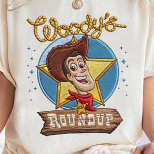 Può includere: T-shirt color crema con un design ricamato di Woody di Toy Story. Il design include il volto di Woody, un distintivo da sceriffo e le parole "Woody's Roundup" in un font in stile western. Il design è ambientato su un cerchio blu e una stella d'oro.