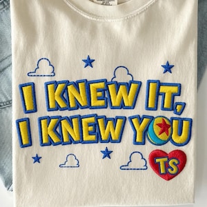 May include: An off-white t-shirt featuring embroidered text that reads I KNEW IT, I KNEW YOU in yellow letters with blue outlines. The design includes blue embroidered clouds, stars, a colorful ball icon, and a red heart containing the yellow letters TS.