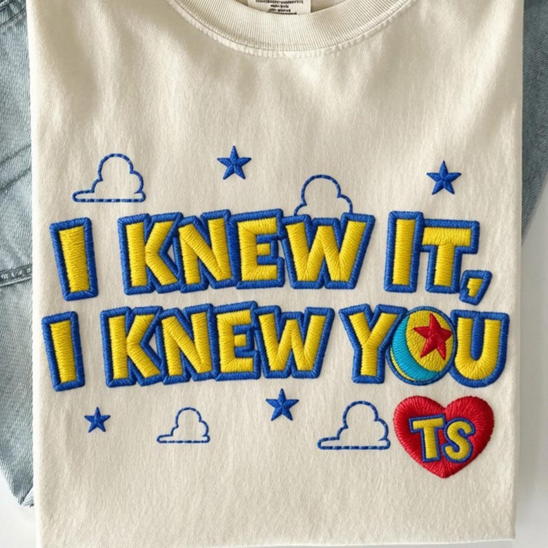 May include: An off-white t-shirt featuring embroidered text that reads I KNEW IT, I KNEW YOU in yellow letters with blue outlines. The design includes blue embroidered clouds, stars, a colorful ball icon, and a red heart containing the yellow letters TS.
