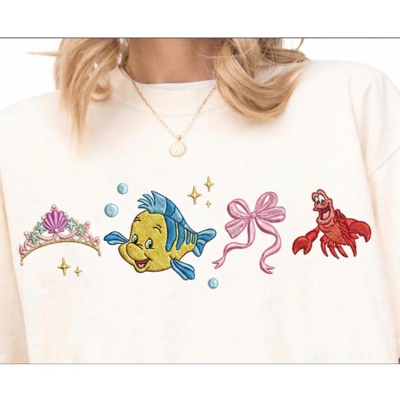 Puede incluir: Sudadera color crema con dise&ntilde;os bordados de una tiara, un pez amarillo y azul, un lazo rosa y un cangrejo rojo. El dise&ntilde;o incluye burbujas azules y estrellas doradas.