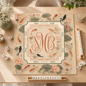 May include: A square mahjong mat featuring a beige background with a central red monogram of the letters SMB. The border is decorated with illustrations of orange, red, and black koi fish swimming among pink lotus flowers and green lily pads.