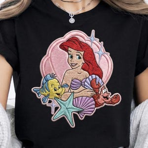 Pode incluir: Camiseta preta com um design bordado de Ariel de A Pequena Sereia. O design inclui Ariel, Linguado, Sebastião, uma estrela do mar e uma concha, tudo em um fundo rosa com detalhes de estrelas azuis.