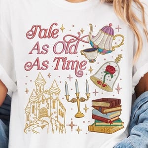 Puede incluir: Camiseta blanca con un diseño bordado con el texto "Tale As Old As Time". El diseño incluye un castillo, una tetera, una taza de té, libros, una rosa en una campana y un candelabro. El diseño es en tonos rosa, dorado y azul.