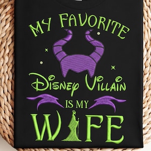 Op de afbeelding: Zwart T-shirt met groen en paars geborduurde tekst en afbeeldingen. De tekst luidt: "MY FAVORITE Disney Villain IS MY WIFE." Het ontwerp bevat een paarse gehoornde hoofdtooi en een groen silhouet van een personage.