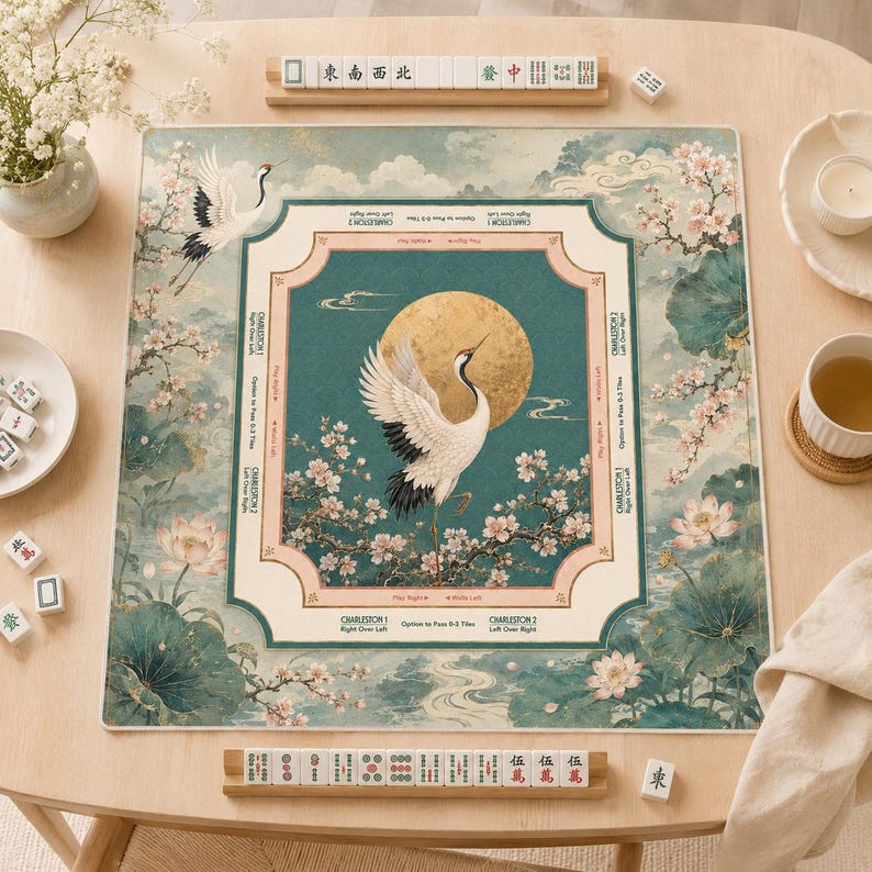 Op de afbeelding: Een vierkante Mahjong-mat met een centrale gouden maan, een witte kraanvogel en kersenbloesems op een groenblauwe achtergrond. De rand bevat gedrukte instructies voor de Charleston-spel fase. Witte Mahjong-tegels liggen op houten rekjes.