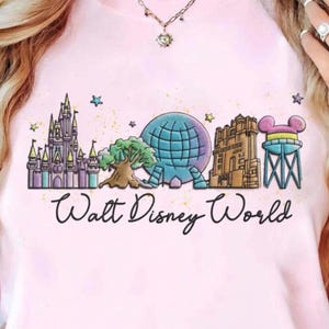 May include: Pale pink t-shirt with a graphic of Walt Disney World landmarks, including a castle, Spaceship Earth, and a water tower with Mickey Mouse ears. The words "Walt Disney World" are written in cursive below.