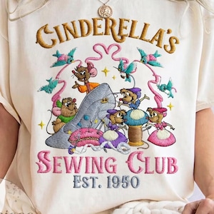 Puede incluir: Camiseta color crema con un diseño bordado del Club de Costura de Cenicienta. El diseño incluye la zapatilla de cristal de Cenicienta, ratones, pájaros y el texto "CINDERELLA'S SEWING CLUB EST. 1950".