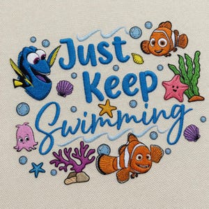 Puede incluir: Diseño bordado con la frase "Just Keep Swimming" en escritura azul, rodeado de criaturas marinas de dibujos animados. Incluye un pez cirujano azul, un pez payaso, una estrella de mar y burbujas sobre un fondo beige. El diseño tiene un tema submarino caprichoso.