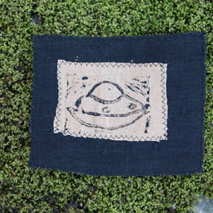 Pode incluir: Um remendo de tecido preto com um quadrado de tecido bege costurado no topo. O quadrado bege tem uma imagem em preto e branco de um disco voador.