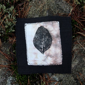 Può includere: Un quadrato di tessuto nero con un quadrato di tessuto bianco all'interno. Il quadrato di tessuto bianco ha una stampa di foglia nera al centro.