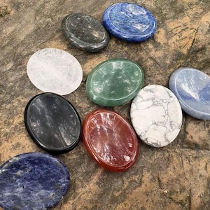 May include: A collection of polished, oval-shaped gemstones in various colors, including blue, green, red, black, and white. The stones are displayed on a textured, brown surface, showcasing their natural patterns and textures.