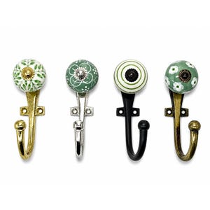 Peut inclure: Quatre patères murales avec des boutons en céramique ronds aux motifs variés. La première a une base dorée et des feuilles vertes, la deuxième une base argentée avec des motifs floraux, la troisième une base noire à rayures vertes et la quatrième une base bronze avec des fleurs blanches.